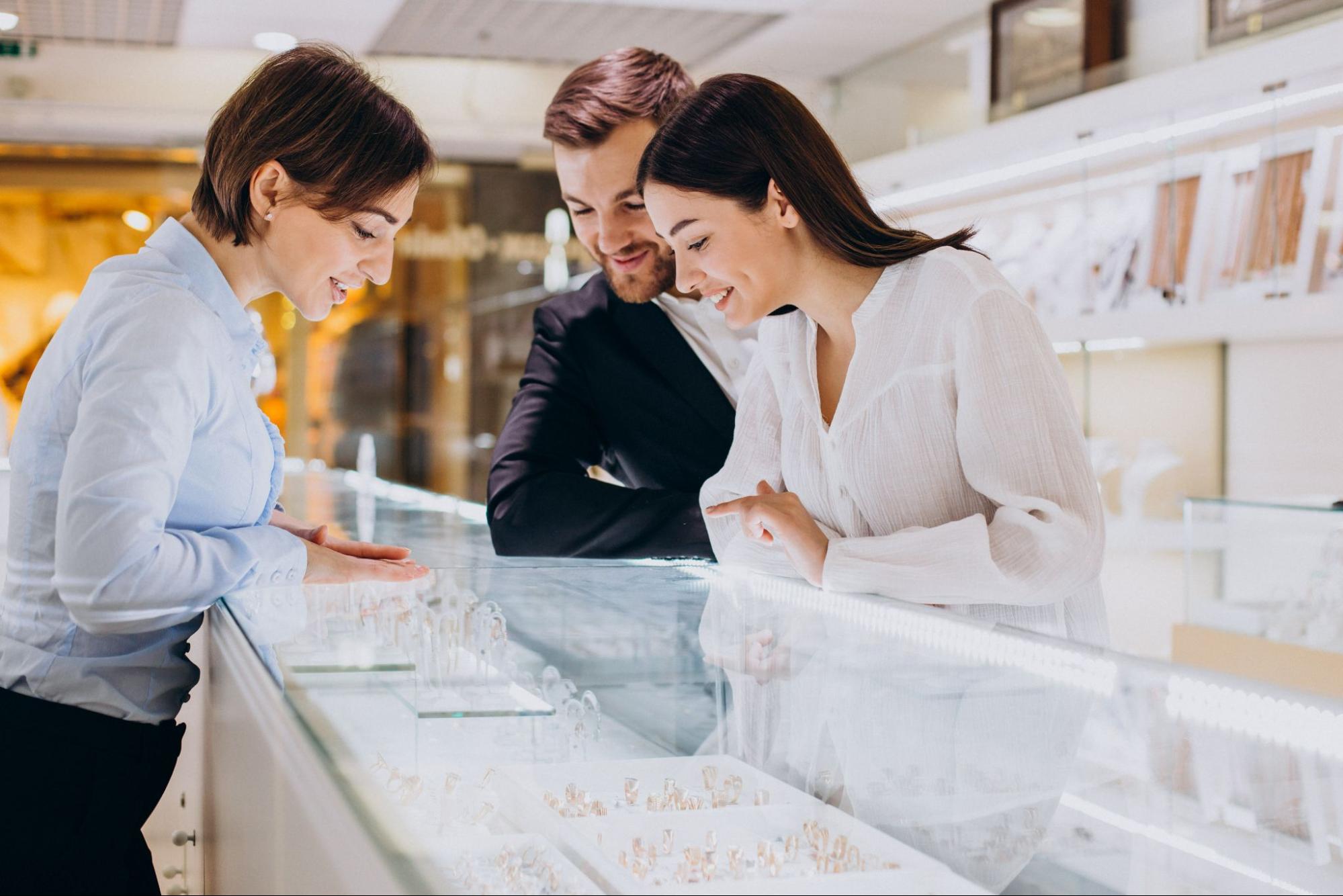 A couple stands behind a jewelry counter and taks to a jewelry as the shop for engagement rings together